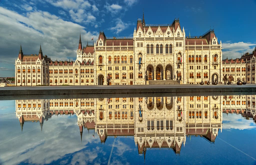 The majestic Hungarian Parliament Building in Budapest reflected perfectly in a still pool of water under a cloudy blue sky.