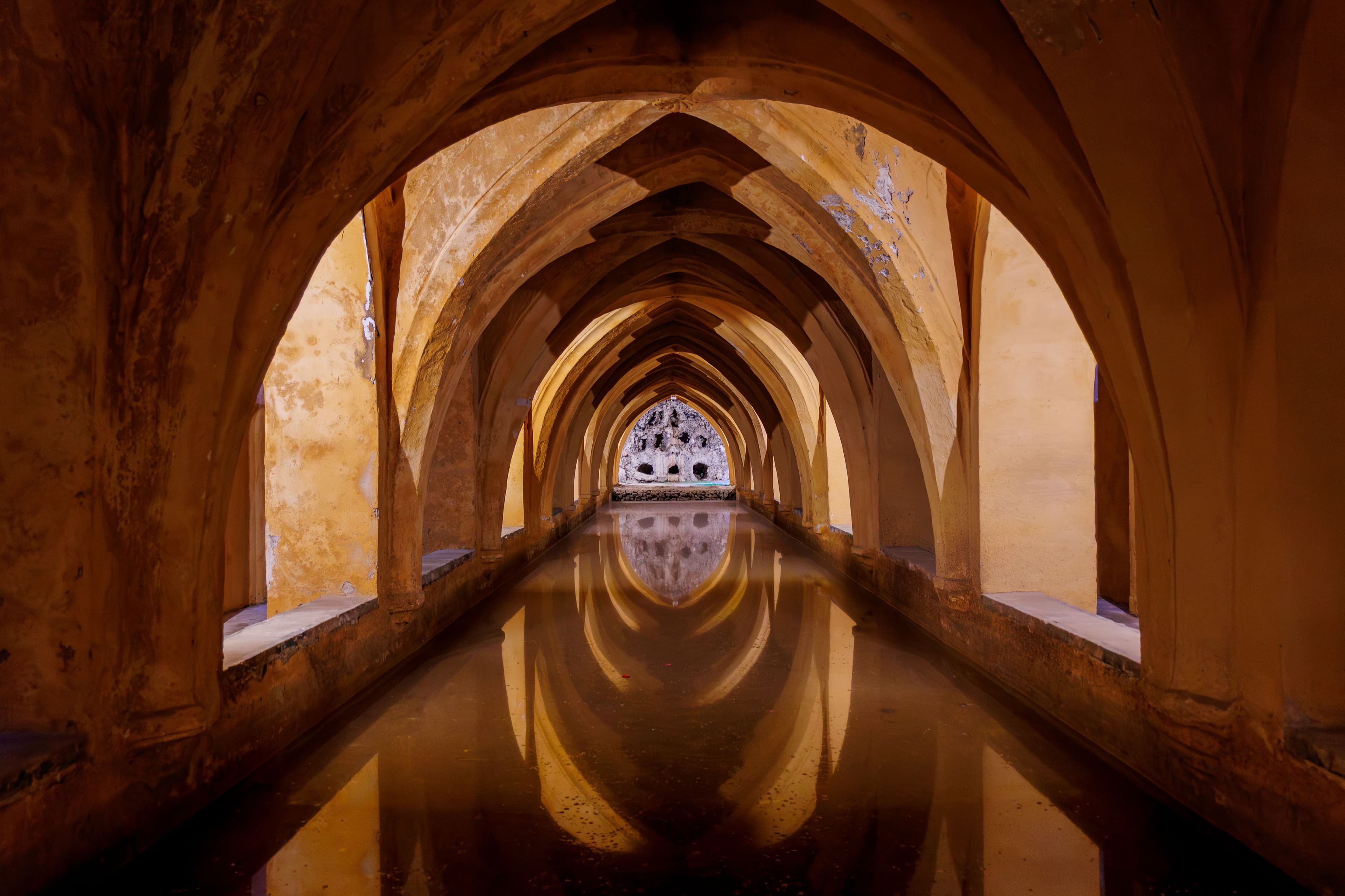 Symmetrical vaulted arches reflecting in the still water of the Baños de Doña María at the Royal Alcázar of Seville.