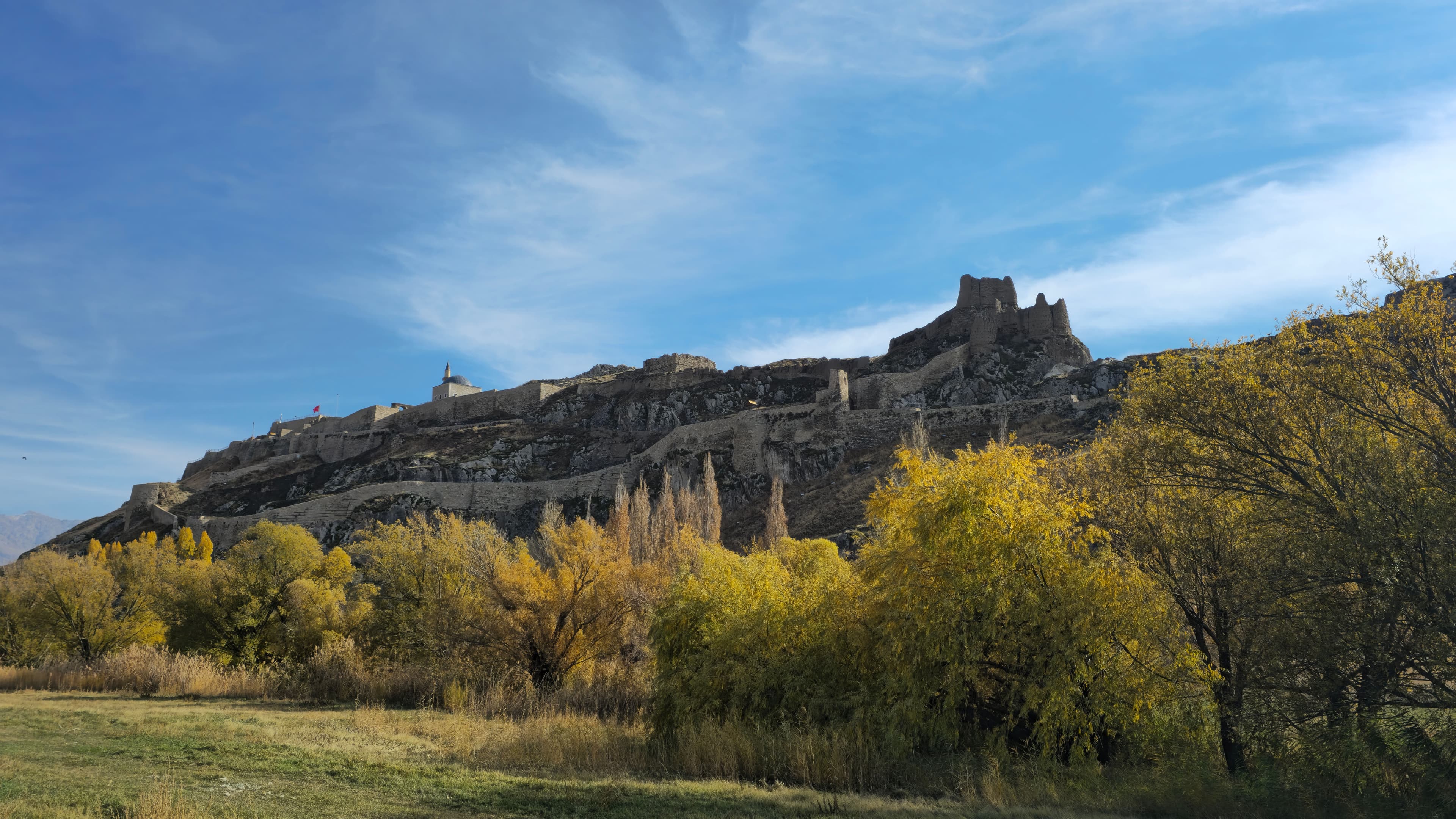 A panoramic view of the ancient Van Castle ruins atop a rocky hill surrounded by autumn trees in Turkey.